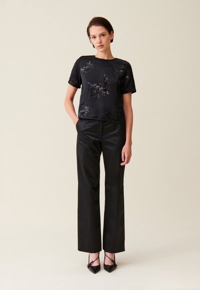 Top in satin nero con ricami floreali, abbinato a pantaloni neri a vita alta e décolleté neri a punta. Design semplice ed elegante.