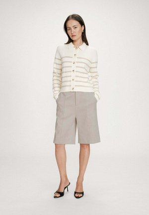 Femme debout, mains dans les poches, portant un cardigan à rayures blanches et beiges boutonné, un short beige mi-cuisses et des sandales à talons noires.