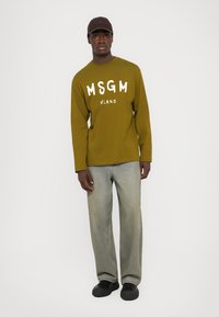 Pikksiniste varrukatega oliivroheline T-särk, millel on valge "MSGM Milano" tekst, koos heledate denimpükstega ja mustade kingadega. Pruun müts lõpetab välimuse.
