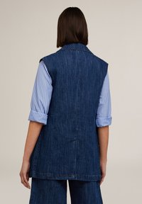 Tummansininen farkkutakki ilman hihoja, jossa on näkyvät ompeleet, korkea kaulus ja yksinkertainen takasuunnittelu, yhdistettynä vaaleansiniseen raidalliseen paitaan.