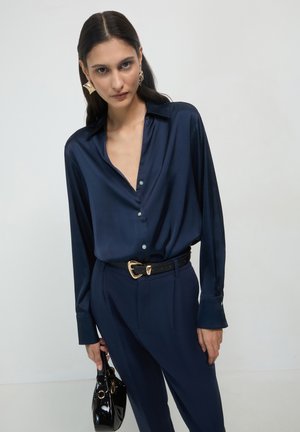 Woman wearing navy silk blouse and pants, holding small black patent handbag, with gold earrings and gold-buckled belt, against plain background.