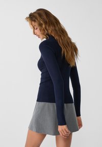 Marineblauer, taillierter Pullover mit langen Ärmeln und geripptem Saum, kombiniert mit einem hellgrauen, plissierten Rock mit feinen vertikalen Streifen.