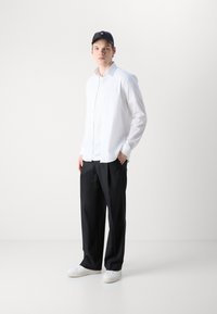 Weißes Hemd mit langen Ärmeln und lockerer Passform, kombiniert mit schwarzen weiten Hosen und weißen Sneakers. Eine schwarze Mütze rundet das Outfit ab.