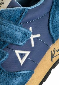 Primo piano di una sneaker blu con dettagli in tessuto testurizzato, triangolo bianco cucito e simboli più, e suola intermedia beige con bordo nero.