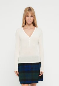 Λευκό μακρυμάνικο τοπ με V-λαιμόκοψη και πλάκα με τρία κουμπιά. Συνδυάζεται με μαύρη και μπλε φουστάνια καρώ. Ομαλή υφή, εφαρμοστή σχεδίαση.