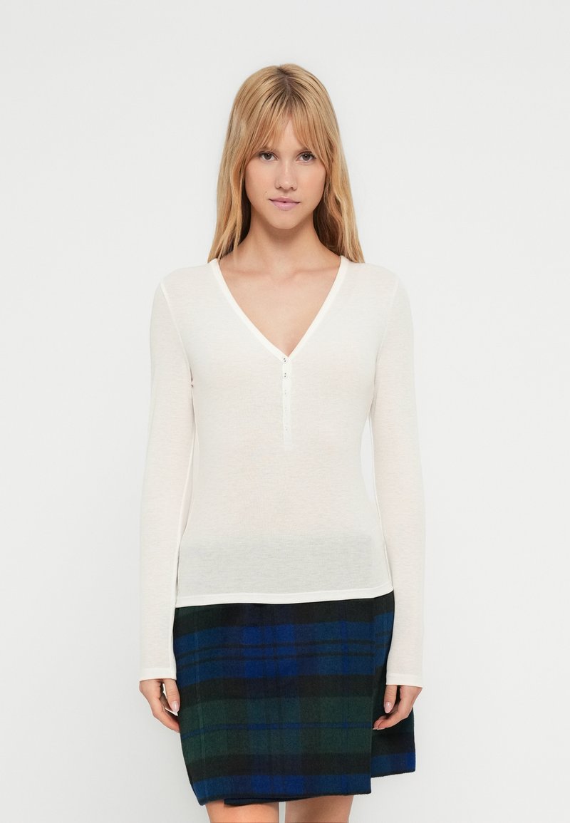 Λευκό μακρυμάνικο τοπ με V-λαιμόκοψη και πλάκα με τρία κουμπιά. Συνδυάζεται με μαύρη και μπλε φουστάνια καρώ. Ομαλή υφή, εφαρμοστή σχεδίαση.