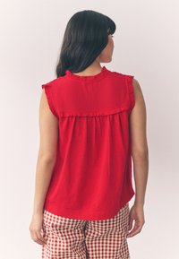 Rotes ärmelloses Oberteil mit strukturierter Stoff, Rüschenkragen und plissiertem Rückendesign; kombiniert mit karierten Hosen in Rot und Weiß.