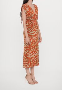 Femme portant une robe midi à motifs orange et beige avec des détails froncés et des bretelles à nouer, associée à des sandales à talons beiges.