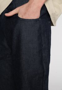 Jean en denim foncé avec une texture lisse, des coutures visibles et un détail de poche avant. Le tissu a un léger éclat sur un fond clair.