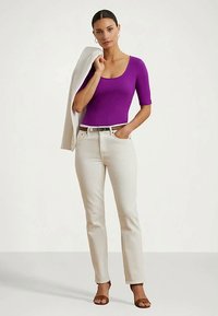 Γυναίκα με μωβ τοπ και μπεζ τζιν, κρατώντας ένα μπεζ μπουφάν στον έναν ώμο, να στέκεται μπροστά σε λευκό φόντο.