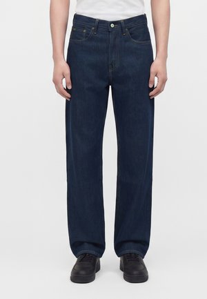 Person iført mørkeblå jeans med lige ben, hvid t-shirt og sorte sneakers, stående mod en ensfarvet lys baggrund.