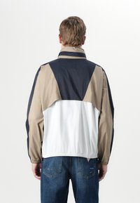 Fargeblokkert vindjakke med paneler i beige, marineblå og hvit, elastisk kant, høy krage og en lett, glatt tekstur.