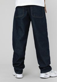 Pantalon en denim bleu foncé à coupe décontractée, avec deux poches arrière et des coutures jaunes contrastantes, associé à des baskets blanches.