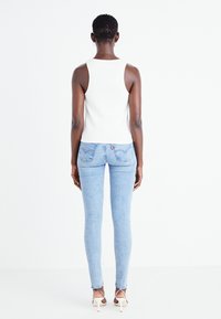 Top sin mangas blanco acanalado, jeans skinny azul claro, tacones con pulsera al tobillo. Textura suave, diseño ajustado y bolsillos traseros detallados con el logotipo de la marca.
