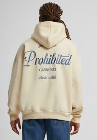 Kremos spalvos, laisvo kirpimo hoodie, kuriame yra mėlyna siuvinėta užrašas "Draudžiami drabužiai" ir "Berlyno-Mitte" nugaros pusėje.