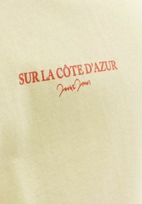 Texte rouge « SUR LA CÔTE D'AZUR » et une signature rouge imprimés sur un tissu jaune clair, probablement un gros plan d'un vêtement.