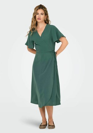 Green wrap dress in textured fabric with short flutter sleeves, a v-neckline, and a side tie, falling to mid-calf, paired with flat shoes.