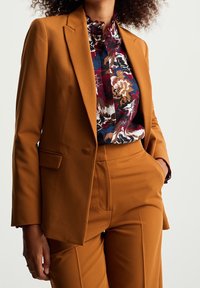 Senapsgul skräddarsydd blazer med knapphålskrage, kombinerad med en mörkt blommig blus och matchande hög midja byxor.