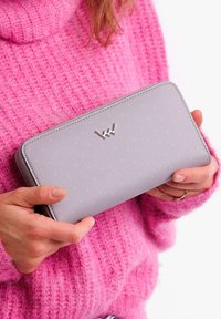 Gray wallet with pink polka dots, smooth texture, logo accent on front, zip closure, held in hands, wearing a pink knit sweater.