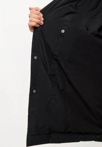 Interior de chaqueta negra que muestra una textura suave, botones a presión y costuras de bolsillos ocultos. Diseño minimalista con una forma elegante y aerodinámica.