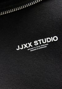 Sudadera negra con una textura suave que presenta texto impreso en blanco: "JJXX STUDIO" y otra marca adicional. Cierre de cremallera completa visible.
