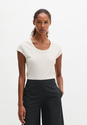 Femme aux cheveux bouclés attachés porte un haut blanc côtelé à manches courtes et un pantalon noir taille haute, debout devant un fond uni.