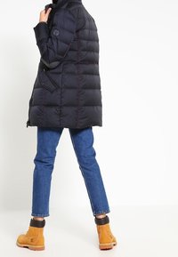 Zwarte donsjack met doorgestikt ontwerp, hoge kraag en zijzakken. Draagt blauwe spijkerbroek en beige enkellaarzen met zwarte accenten.