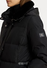 Melnā pūkainā jaka ar mīkstu, mākslīgās kažokādas apkakli, sudrabīgiem aksesuāriem un logotipa uzšuvumu uz piedurknes. Gluda auduma struktūra ar šūtu daļu.