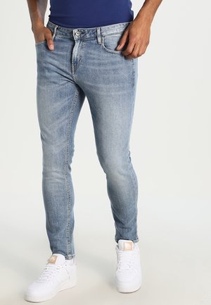 Man draagt strakke lichtblauwe spijkerbroeken en witte sneakers, met handen in de zakken, staand tegen een effen lichte achtergrond.