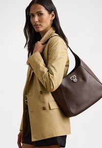 Brown leather shoulder bag with rounded shape, featuring silver hardware and a decorative clasp, held by a figure in a beige blazer.