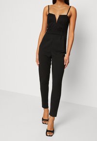 Mono negro con cuerpo de encaje, escote pronunciado y tirantes finos. Los pantalones ajustados se estrechan en el tobillo. Lleva unos tacones negros destalonados.