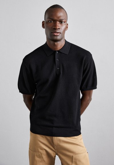 Sort polo-shirt med korte ærmer, tekstureret stof, to-knaps placket og krave. Bæres med lyse bukser.