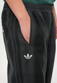 Melnas sporta bikses ar skotu rakstu, elastīgu jostasvietu, sānu kabatām un baltu izšūtu Adidas logo kreisajā augšstilbā.