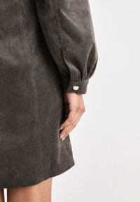 Tmavozelené kordové šaty s naťahovacími rukávmi, zapínaním na gombíky na manžetách a hladkou textúrou so zvislým ryhovaním.