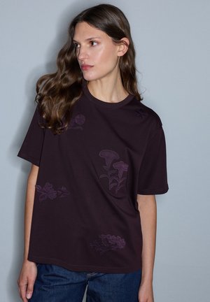 Frau mit langen, welligen braunen Haaren, die ein dunkelviolettes T-Shirt mit subtiler Blumenstickerei und blaue Jeans vor einem schlichten grauen Hintergrund trägt.
