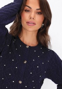 Laivasto sininen kardigan, jossa on teksturoitu neulekuvio, koristeltu hopeanvärisillä niiteillä ja kultasävyisillä napilla, jossa on pyöreä pääntie.