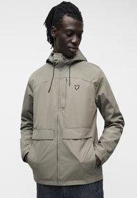 Homme à la peau foncée et aux dreadlocks portant une veste à capuche olive clair avec deux poches avant et un logo d'oiseau jaune sur la poitrine.