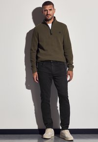 Suéter verde oliva con cuello de media cremallera, jeans de mezclilla negros y zapatillas en tonos blanco roto. Textura suave con un detalle de logo minimalista.