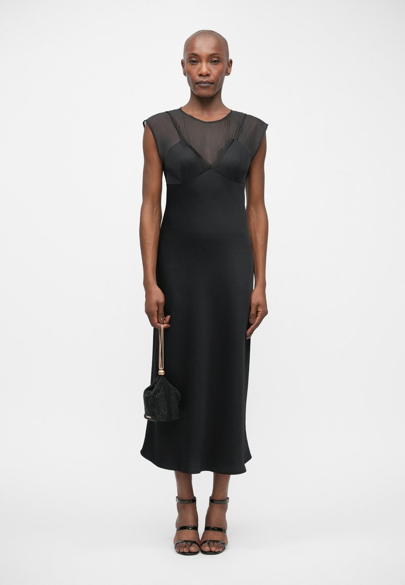 Black midi dress with sheer mesh bodice, fitted silhouette, and cap sleeves. Model holds a small black embellished handbag and wears strappy heels.