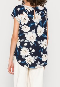 Květinová námořnická blůza s krátkými rukávy, zdobená bílými a světle růžovými květinami, modrými listy a detaily ve tvaru klíčové dírky na zádech. Měkká tkanina s kulatým spodním okrajem.