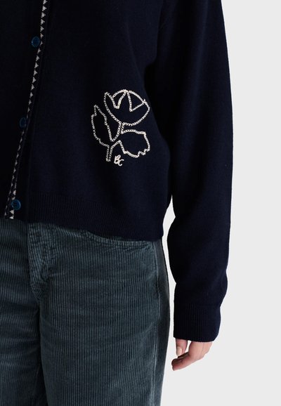 Personne portant un cardigan bleu foncé avec une broderie florale blanche et des boutons bleus, associé à un pantalon en velours côtelé gris-vert.