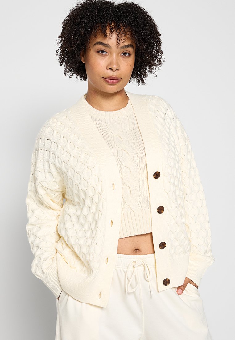 Crème-kleurige gebreide cardigan met een gestructureerd ruitpatroon, vijf bruine knopen en geribbelde manchetten. Draag het over een lichte gebreide top.