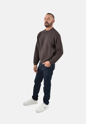 Maglione a coste marrone scuro, jeans blu e sneakers bianche. Il modello sta in piedi con una mano in tasca, presentando una vestibilità rilassata e un design moderno.