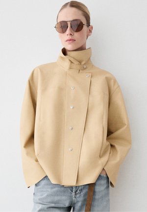 Femme portant une veste beige à col montant avec une fermeture asymétrique à boutons et des lunettes de soleil, mains dans les poches d'un jean bleu clair.