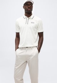 Άνδρας με λευκό πόλο πουκάμισο με μαύρο περίγραμμα και ανοιχτό μπεζ παντελόνι, με τα χέρια στις τσέπες, κοιτάζει ελαφρώς προς τα πλάγια, φοράει λευκό καπέλο.