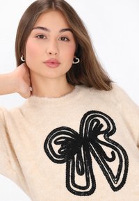 Beige tröja med en svart broderad fjärilsdesign; mjuk textur, rund halsringning och subtil ribbning vid ärmslut och fåll.