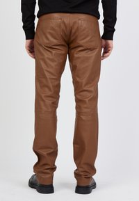 Braune Lederhosen mit glatter Textur, tailliertem Design und zwei Gesäßtaschen, kombiniert mit schwarzen Schuhen.