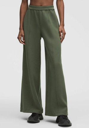 Persona che indossa pantaloni a vita alta, a gamba larga color verde oliva, abbinati a scarpe sportive nere, in piedi su uno sfondo semplice e chiaro.