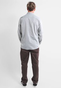 Chemise à manches longues gris clair, à boutons, avec une texture lisse et un détail au dos, associée à un pantalon marron foncé et des chaussures noires.
