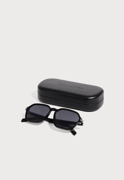 Lunettes de soleil géométriques noires avec verres foncés à côté d’un étui rigide noir de la marque Marc Jacobs sur fond blanc.